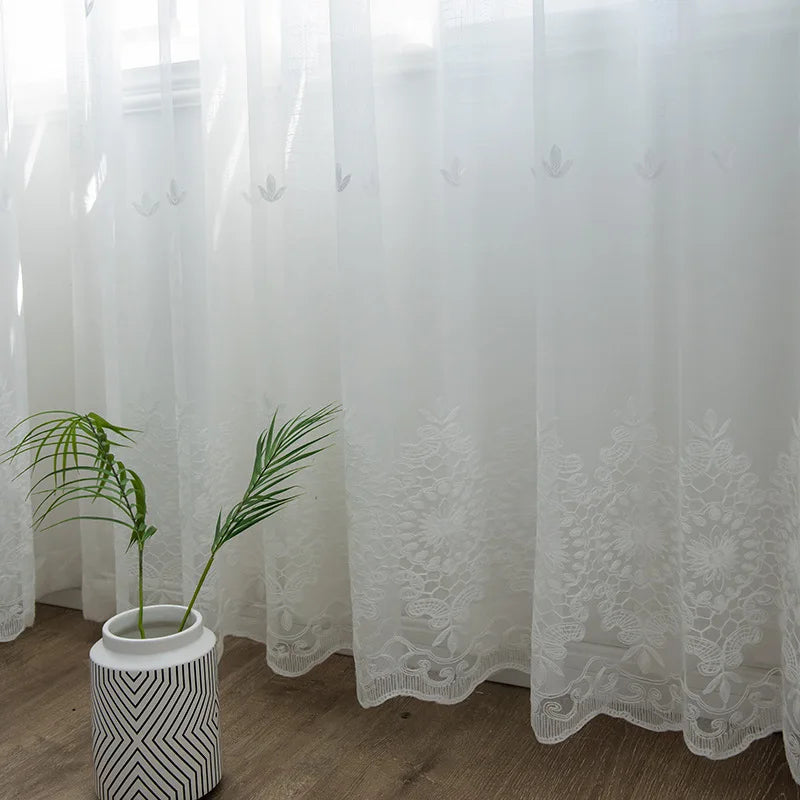 Bas d’un voilage blanc transparent brodé à motifs floraux, avec une plante verte posée dans un pot moderne à rayures noires et blanches.