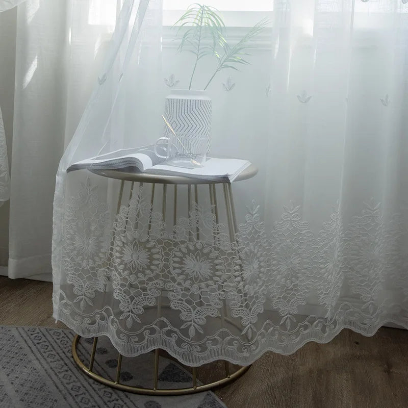 Zoom sur un voilage blanc en 140x200 posé devant une fenêtre, laissant apparaître un vase et un livre sur une table en métal doré, avec broderies florales apparentes.
