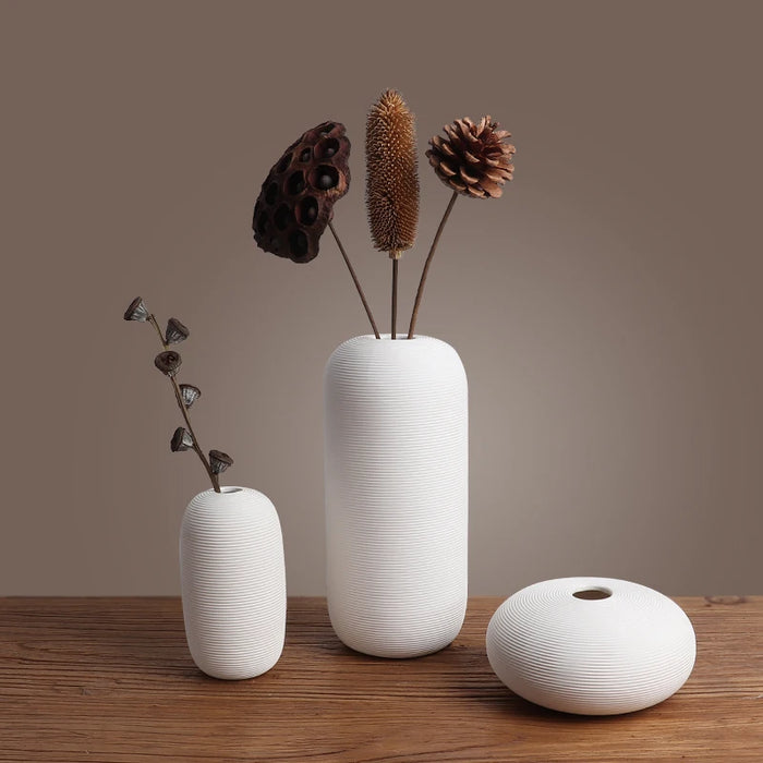 Plusieurs vases blancs rayés avec des fleurs sèches brunes posés sur une table en bois.