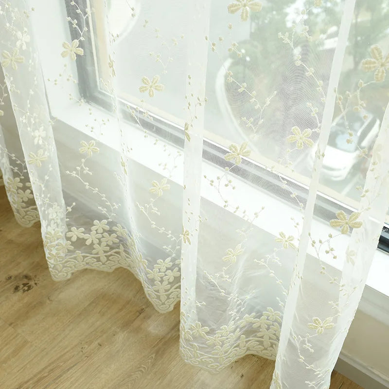 Voilage brodé floral installé sur une grande baie vitrée, laissant filtrer délicatement la lumière tout en créant un jeu d’ombres poétique.