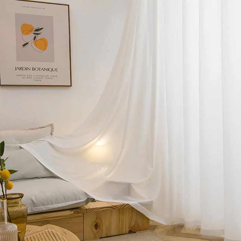 Voilage blanc léger partiellement relevé, posé dans un salon cosy avec banquette basse en bois clair, coussins blancs et lumière tamisée.