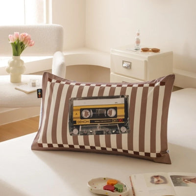 Coussin décoratif imprimé de bandes marron et blanches avec une ancienne cassette audio Denon au centre, placé sur une table blanche dans un intérieur lumineux avec un vase de tulipes roses.