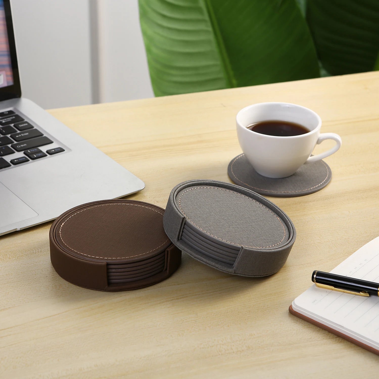 Sur un bureau en bois se trouvent un ordinateur portable, une tasse de café et un ensemble de sous-verres en cuir de différentes couleurs, formant un espace de travail confortable et professionnel.