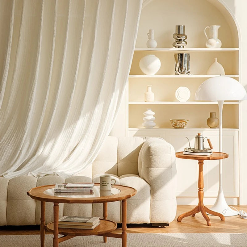 Voilage blanc effet froissé suspendu en arrière-plan d’un salon beige au mobilier organique et aux touches de bois naturel.