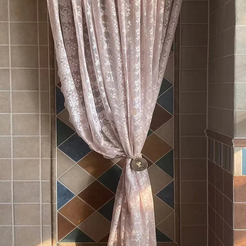 Rideau en dentelle rose transparent noué avec un embrasse, devant un mur carrelé en losanges multicolores pour une déco rustique et colorée.