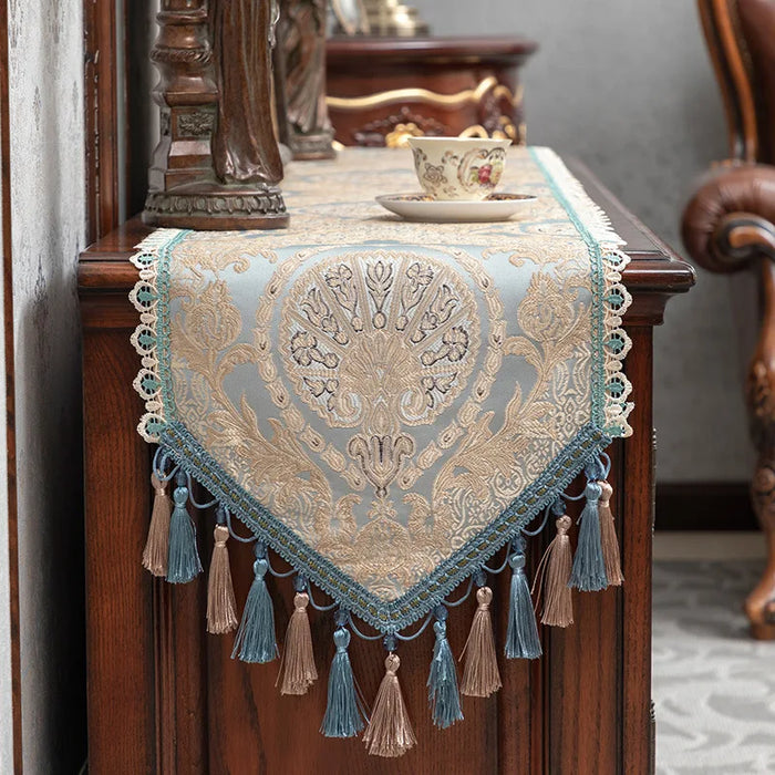Un élégant tapis de table décoratif ornementé de motifs floraux dorés sur un fond bleu clair, entouré de glands et de franges multicolores, créant une atmosphère chaleureuse et raffinée dans cette pièce vintage.