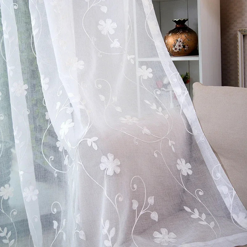 Image d'un rideau de fenêtre blanc en textile brodé avec des motifs roses, utilisé comme traitement de fenêtre pour donner de l'élégance à la pièce. Le même tissu est également utilisé comme nappe de table, créant une harmonie de lin dans la décoration intérieure.