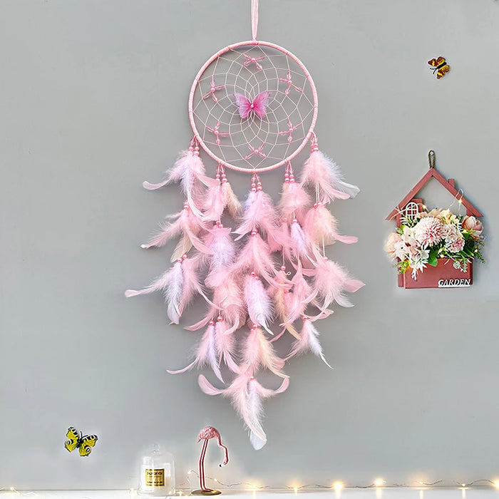 Un attrape-rêves rose en forme de fleur avec des plumes blanches, suspendu sur un mur gris accompagné d'autres décorations.
