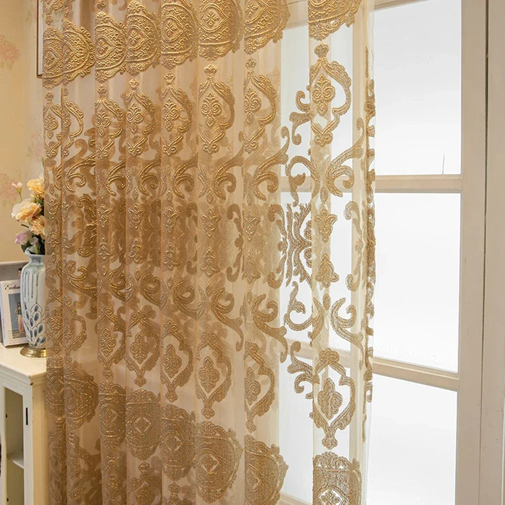 Voilage doré brodé avec motifs floraux orientaux, suspendu devant une fenêtre blanche dans un intérieur lumineux avec meuble classique.