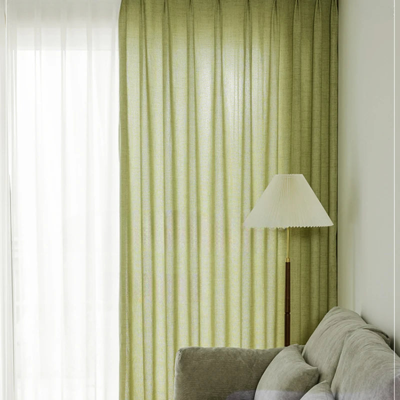 Grand rideau vert amande habillant une baie vitrée, avec un canapé gris et un lampadaire blanc.