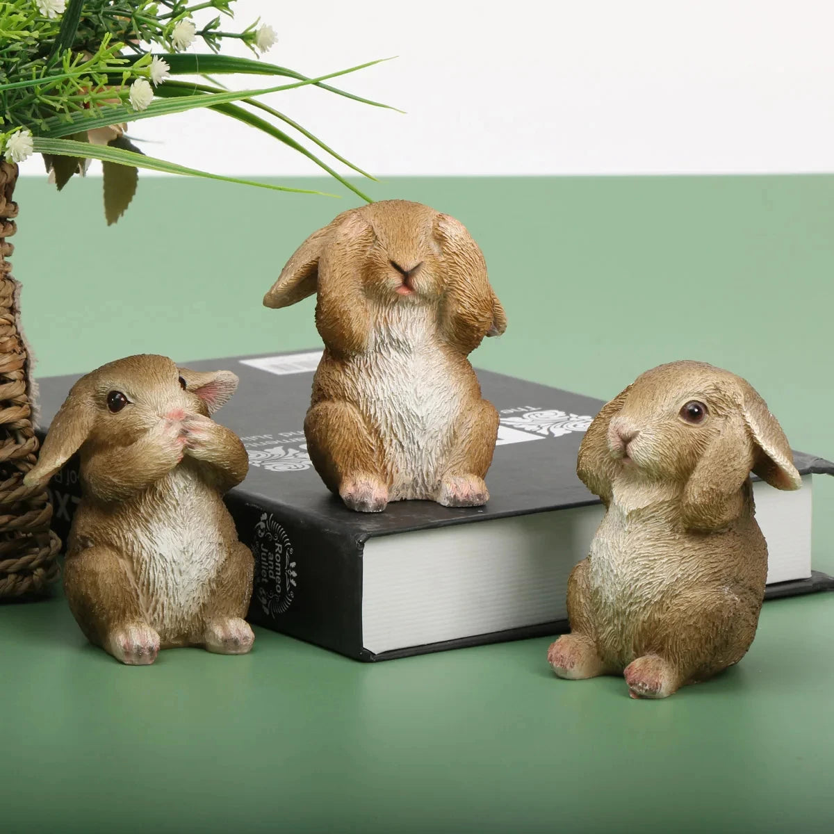 Une famille adorable de lapins en céramique, posée sur un livre avec des plantes verdoyantes en arrière-plan.