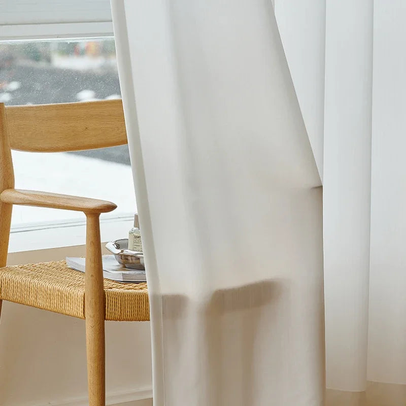 Voilage blanc soufflé par le vent, posé devant une grande fenêtre avec une chaise en bois au style scandinave