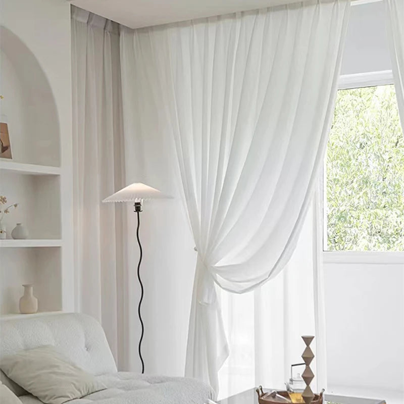 Voilage blanc noué délicatement au centre, dans un salon lumineux avec fauteuil blanc, lampe design noire et vue sur un jardin verdoyant.