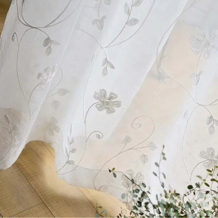 Rideaux blancs avec broderies de géraniums suspendus à une fenêtre, une nappe blanche sur la table en toile de fond et des draps de lin éparpillés autour, créant une ambiance chaleureuse et accueillante. Cette image illustre parfaitement nos gammes de rideaux et de linge de maison aux motifs floraux.