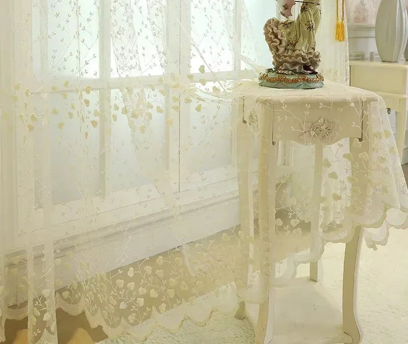 Voilage blanc cassé à fleurs translucides tombant délicatement sur une table d’appoint romantique.
