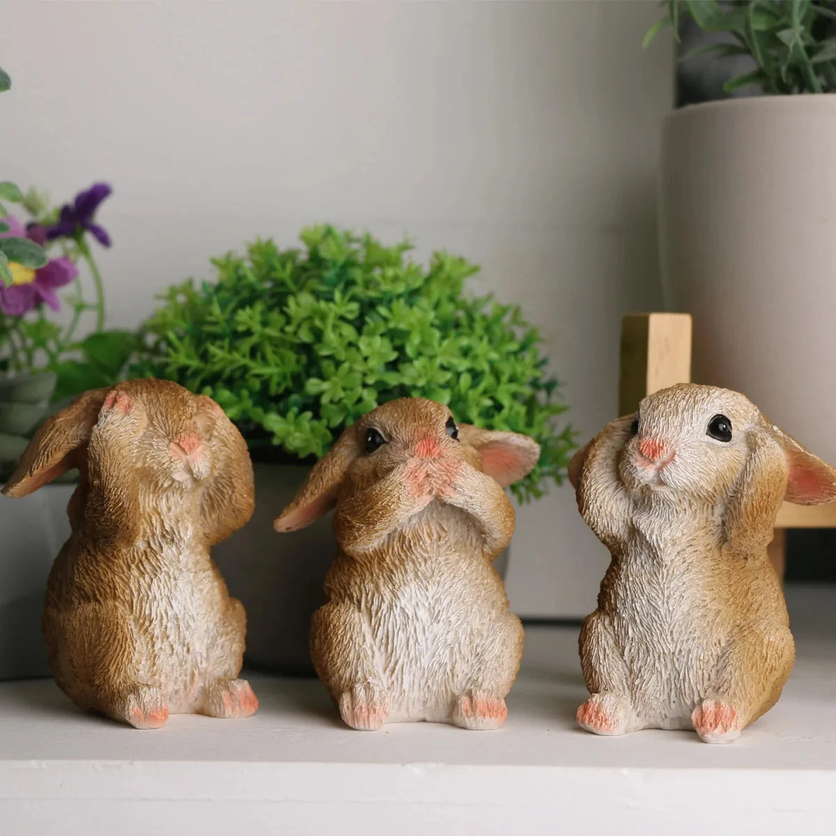 Trois figurines de lapins en porcelaine mignons, assis et regardant vers l'avant, entourés de feuillage vert dans un décor de bureau ou de maison.