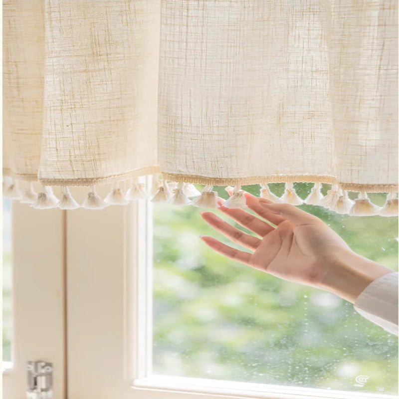 Gros plan sur un rideau crème à pompons avec une main touchant doucement le tissu, devant une fenêtre verdoyante.