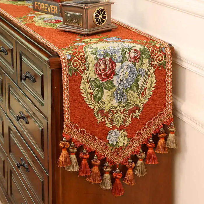 Un magnifique chemin de table brodé de fleurs colorées et de glands décoratifs sur un meuble ancien ajoute une touche élégante et vintage à cette scène.
