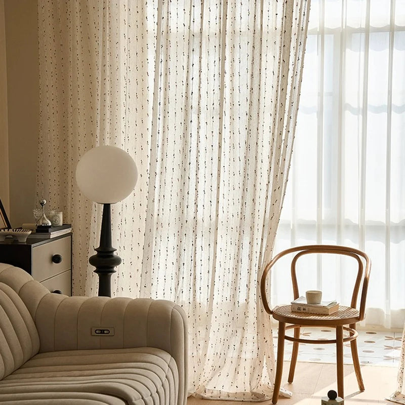 Détail du voilage transparent crème orné de petits pois colorés dans un coin lecture chaleureux avec fauteuil et chaise en rotin.