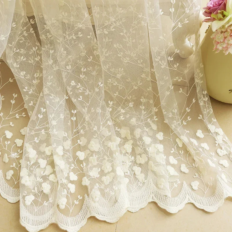 Gros plan sur la broderie florale d’un voilage blanc cassé en dentelle, posé au sol.