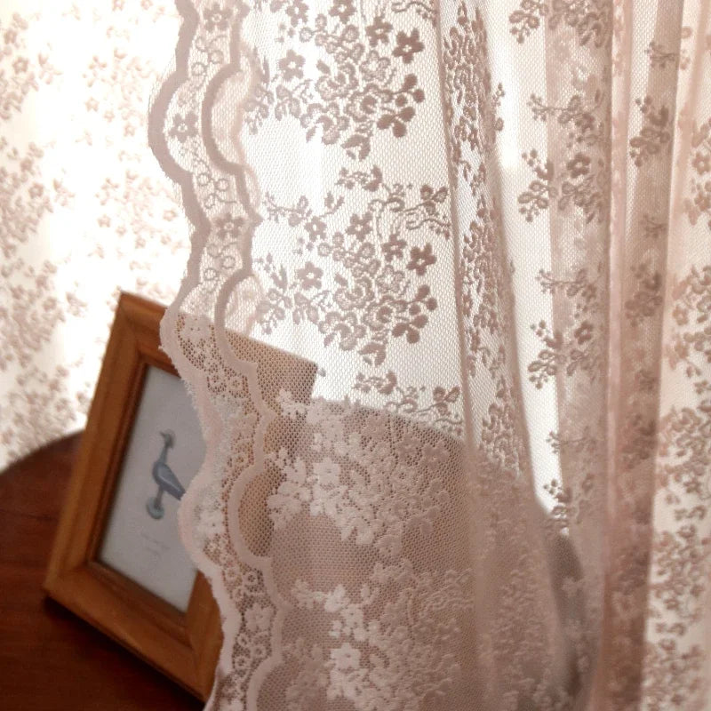 Gros plan sur les bords dentelés et les motifs fleuris du voilage rose pastel, laissant passer doucement la lumière.