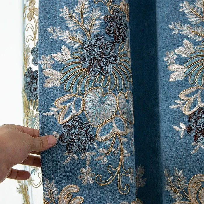 Gros plan sur un rideau bleu pétrole en chenille brodé de motifs floraux dorés et bleu nuit, tenu par une main pour montrer l’épaisseur et la texture du tissu.