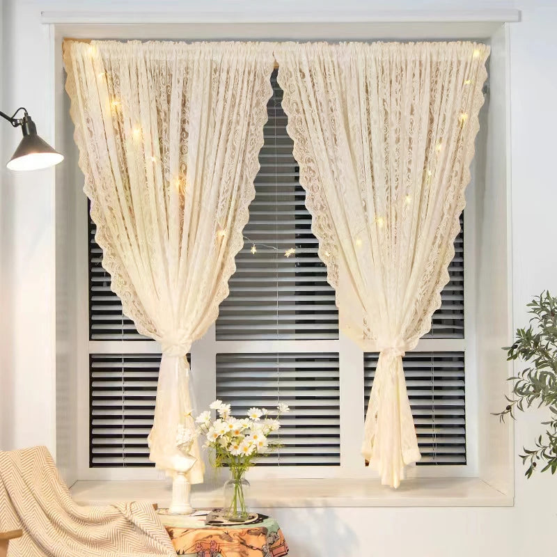 Voilage beige à motif dentelle installé sur une fenêtre à stores horizontaux, agrémenté d'une guirlande lumineuse, dans un intérieur chaleureux avec bouquet de marguerites.