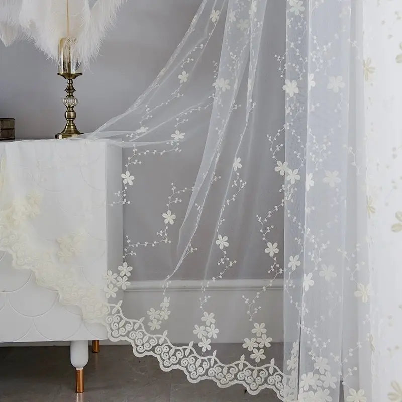 Voilage romantique blanc brodé suspendu sur une fenêtre, accompagné d’un meuble chic et d’un lampadaire plume, pour une ambiance raffinée et féminine.