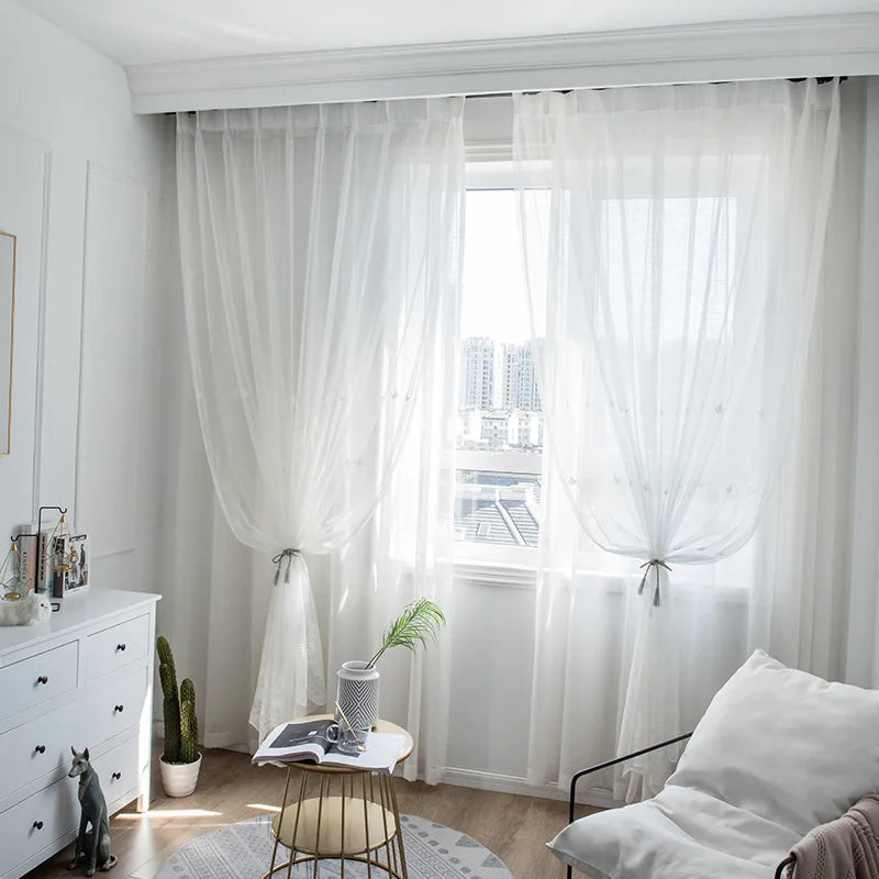 Deux voilages blancs noués avec des embrasses, installés devant une grande fenêtre laissant entrer la lumière naturelle d’un appartement en hauteur.