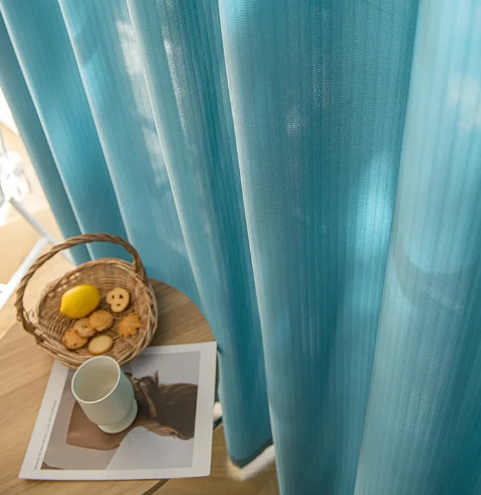 Voilage bleu en 140 x 220 filtrant la lumière naturelle, avec une ambiance cosy grâce à une table en bois, une tasse et un panier de biscuits.
