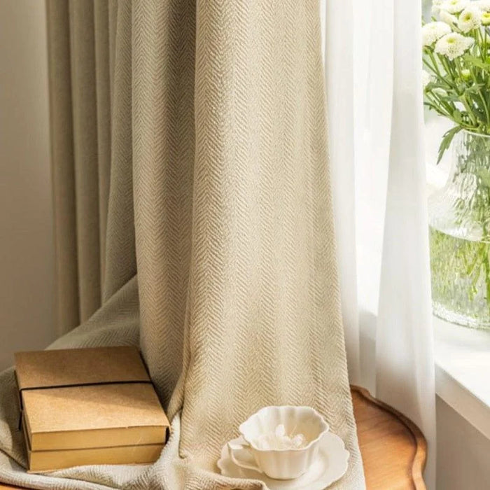 Rideaux élégants couleur beige avec motif chevrons pour salon moderne. Accessoires décoratifs assortis, incluant un vase avec fleurs blanches, posés sur une table en bois éclairée par la lumière naturelle. Ambiance chaleureuse et sophistiquée pour une décoration harmonieuse.