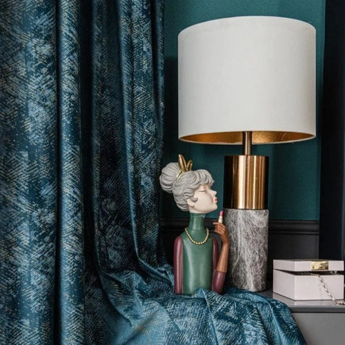 Lampe élégante avec abat-jour blanc et base dorée, posée sur une table d'appoint. À ses côtés, une statuette décorative colorée représente une femme stylisée avec un collier. Rideaux bleu profond avec motifs texturés en arrière-plan, ajoutant une touche de sophistication à l'ensemble du décor. Cette composition raffinée est parfaite pour une décoration intérieure moderne et chic.