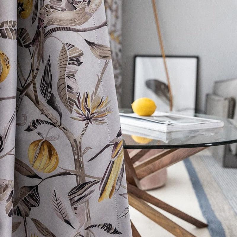 Rideau à motif floral tropical avec des citrons en décoration, suspendu dans une salle à manger contemporaine. Table en verre avec base en bois moderne et un citron posé sur des magazines. Ambiance lumineuse et élégante idéale pour un intérieur raffiné.