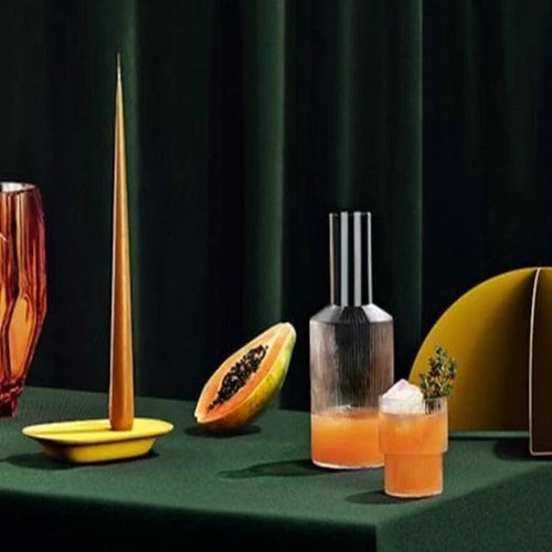 Composition d'un centre de table moderne avec une élégante carafe et un verre contenant une boisson orange. Présentés sur une nappe verte, des éléments décoratifs incluent une bougie effilée jaune, un récipient sculpté et une moitié de papaye. Ce décor élégant et coloré est idéal pour inspirer une atmosphère conviviale et chaleureuse lors d'un dîner ou d'une réception.