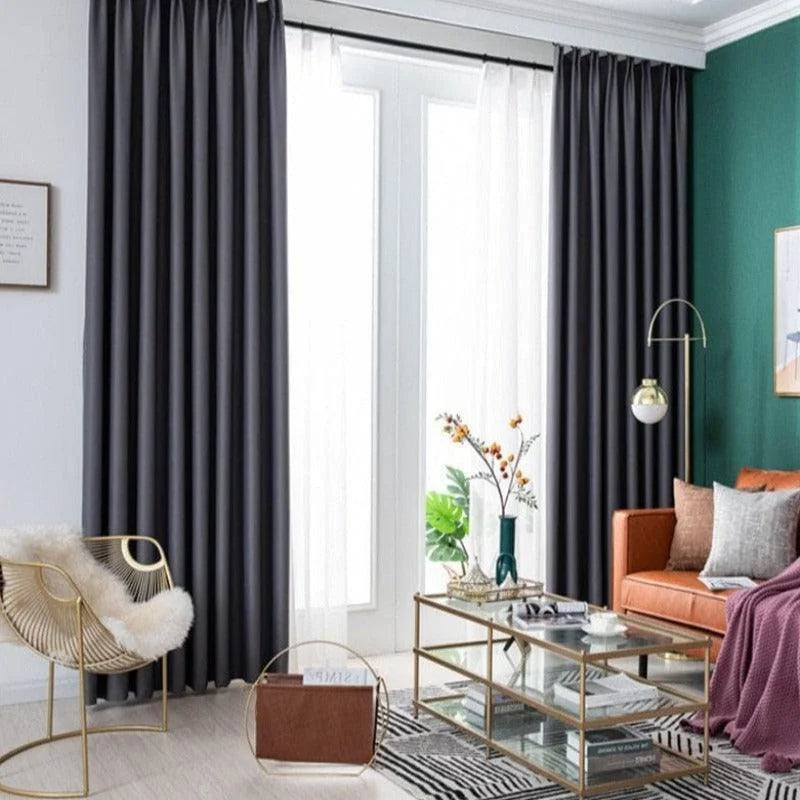 Salle de séjour moderne avec une ambiance élégante, mettant en valeur de longs rideaux de couleur noire qui ajoutent une touche de sophistication. Le canapé orange vif contraste harmonieusement avec le mur vert, créant un espace convivial et accueillant. Une table basse en verre avec des finitions dorées trône au centre de la pièce, entourée d'un fauteuil design et d'éléments décoratifs raffinés. L'éclairage naturel inonde la pièce grâce aux grandes fenêtres, accentuant le