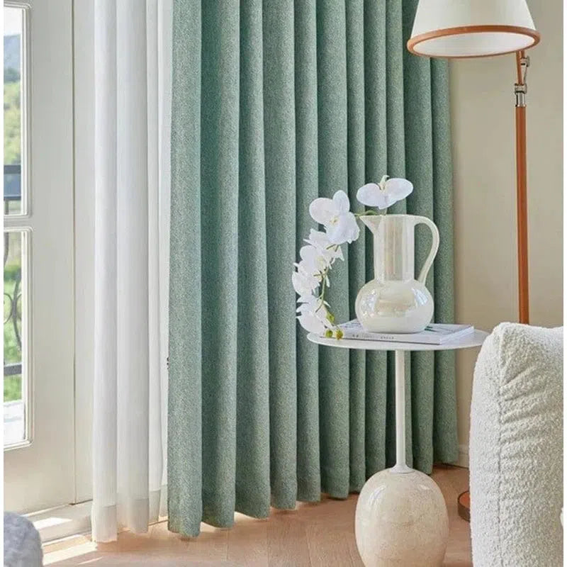 Rideau élégant de couleur bleu-vert, installé dans un salon moderne et lumineux. À côté, une table blanche décorative avec un vase rempli de fleurs blanches, ajoutant une touche florale et raffinée à la pièce. Une lampe sur pied avec un design contemporain complète le décor harmonieux.