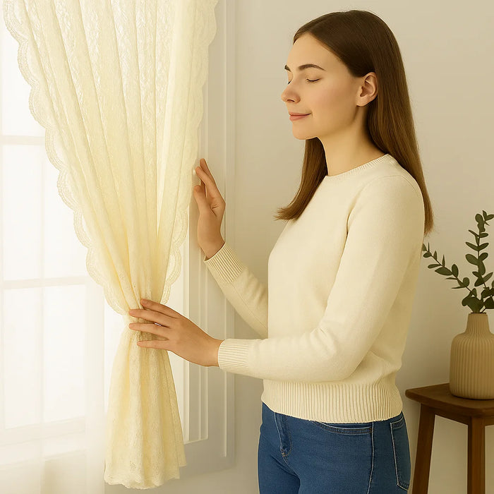 Femme souriante en pull crème touchant un voilage beige clair ajouré, près d'une fenêtre dans une pièce lumineuse à la décoration minimaliste.