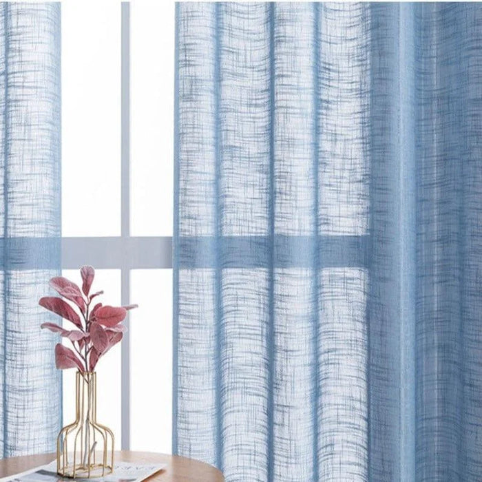 Rideau bleu translucide devant une fenêtre, laissant passer la lumière naturelle, avec une plante décorative dans un vase doré sur une table en bois. Décoration intérieure moderne et élégante.