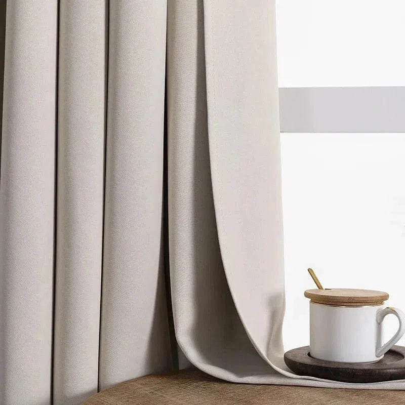 Rideau beige occultant élégant en tissu épais, idéal pour la décoration intérieure moderne. Offre intimité et contrôle de la lumière, parfait pour chambre ou salon. Assorti à une tasse en céramique blanche sur une table en bois clair. Acheter des rideaux de qualité pour un intérieur contemporain et chaleureux.