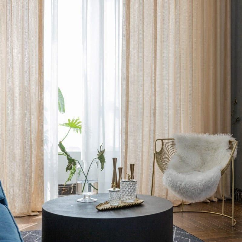Salon moderne avec de longs rideaux beiges encadrant une grande fenêtre, apportant une lumière douce et apaisante. Une chaise design avec un coussin moelleux blanc ajoute une touche de confort. Une table basse noire accueille des accessoires raffinés, créant une ambiance chic et contemporaine. Parfait pour un intérieur élégant et accueillant.