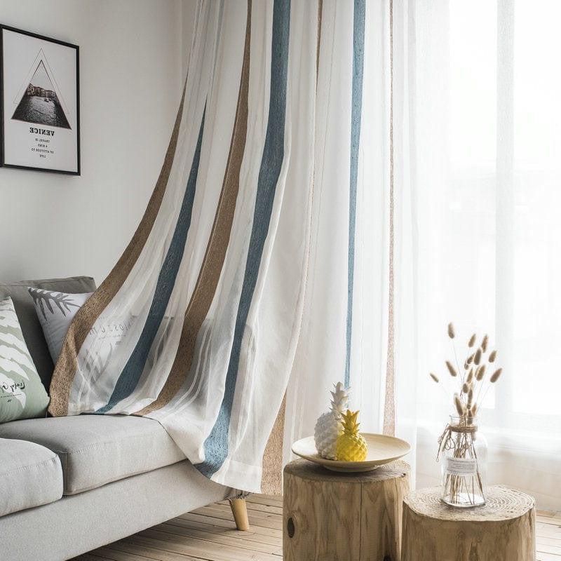 Rideaux modernes à rayures verticales bleues et beiges dans un salon lumineux. Décoration intérieure chic avec canapé gris, coussins à motifs, et table en bois rustique. Accessoires tendance avec ananas décoratif et vase en verre. Ambiance cosy et élégante pour un espace de vie contemporain.