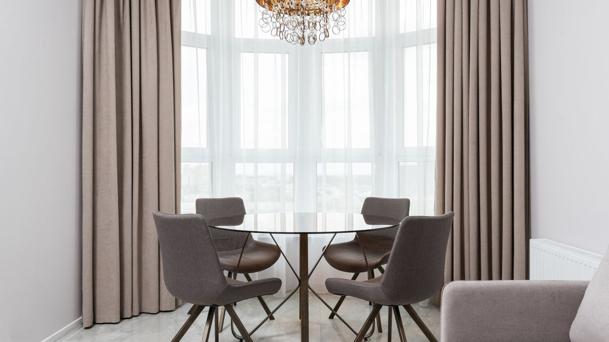 Chaises design grises autour d'une table ronde devant des rideaux taupe et un voilage blanc dans une salle à manger