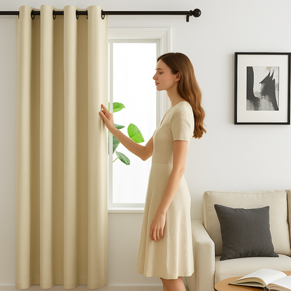 Une femme élégante en robe beige touche un rideau beige dans un salon moderne lumineux avec canapé crème et coussin gris, près d’une fenêtre encadrée de verdure.