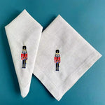 Deux serviettes de table en lin avec des jouets soldats brodés dessus sont posées sur un fond bleu-vert.