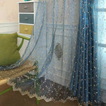Gros plan sur le tissu bleu du voilage orné de broderies florales dorées et blanches, avec mobilier coloré en arrière-plan.