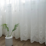 Bas d’un voilage blanc transparent brodé à motifs floraux, avec une plante verte posée dans un pot moderne à rayures noires et blanches.
