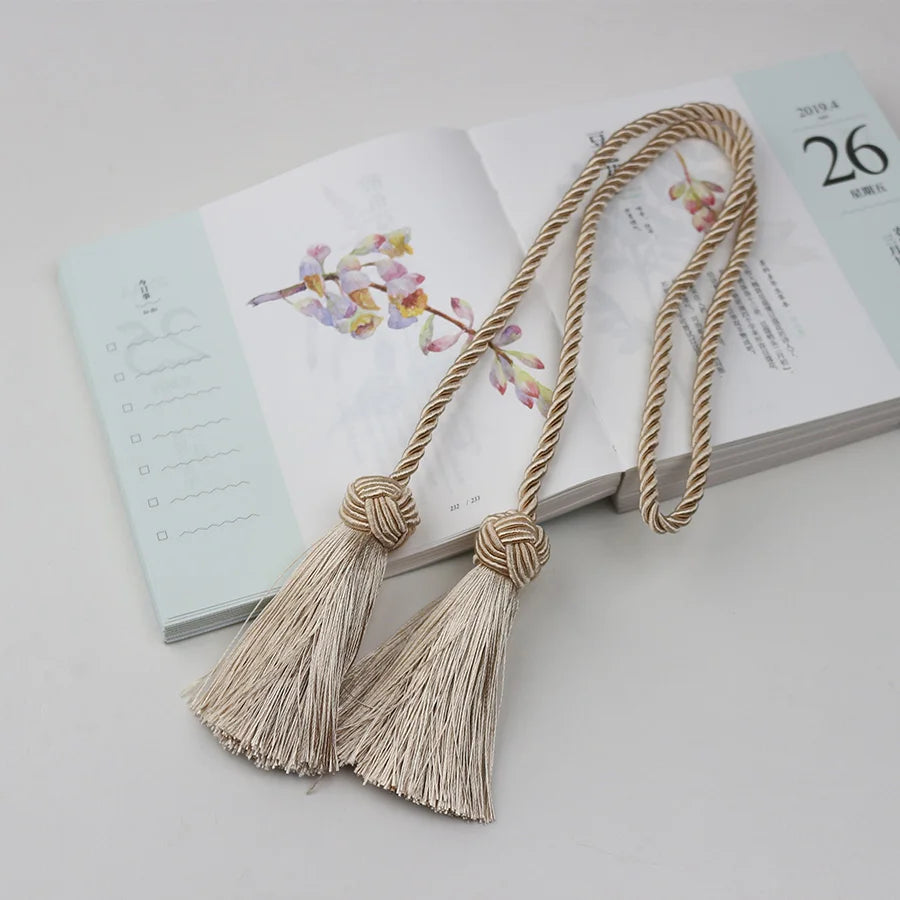 Embrasse rideau torsadée en polyester, finition couleur lin avec deux pompons décoratifs, posée sur un carnet de notes.