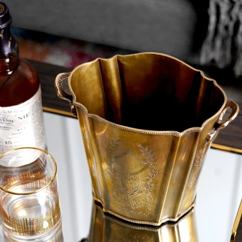 Un vase en métal doré avec des motifs décoratifs en relief, posé près d'une bouteille de vin sur une surface brillante.