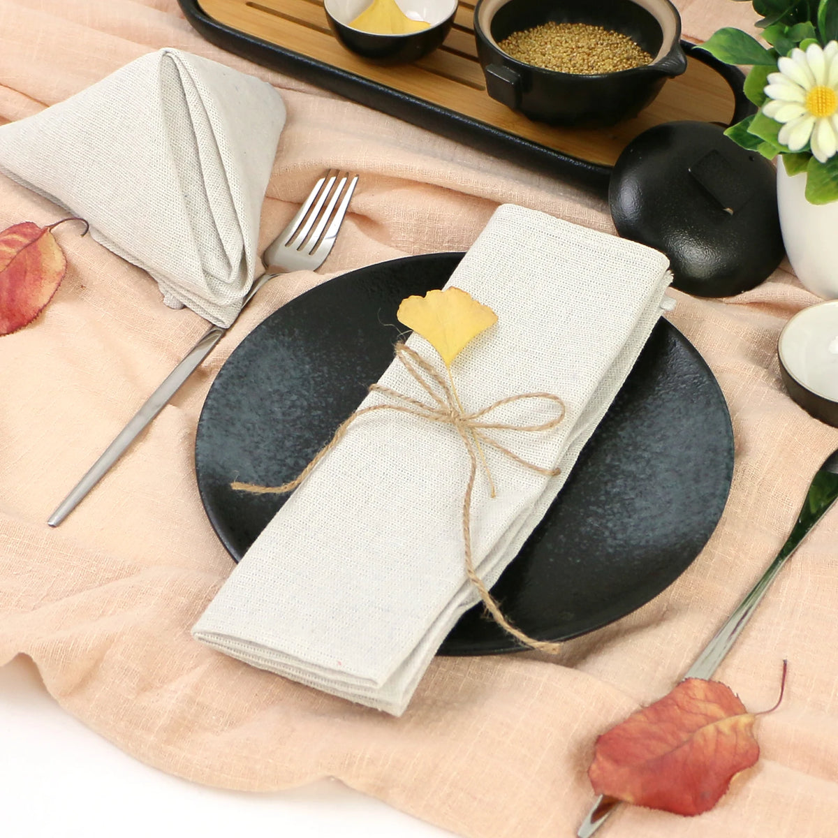 Assiette noire avec couverts, serviette de table blanche, graines et feuille automnale sur plan de travail en bois clair.