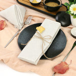 Assiette noire avec couverts, serviette de table blanche, graines et feuille automnale sur plan de travail en bois clair.